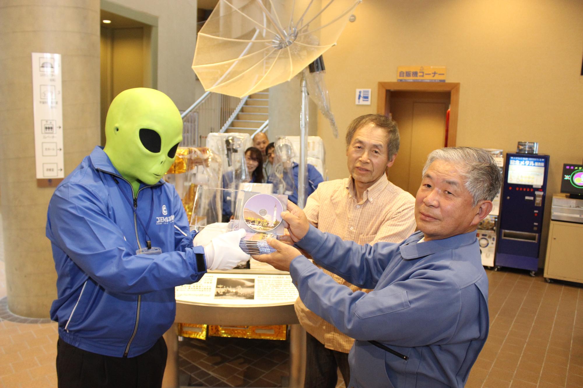 宇宙人サンダーくんにCDを手渡す金井さん（右）と大竹さん（右から2人目）